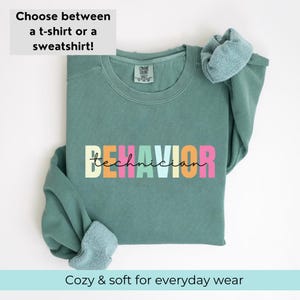 May include: A teal green sweatshirt with the text "BEHAVIOR technician" in rainbow colors. The sweatshirt is folded and displayed on a white background.