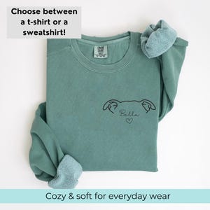 Pode incluir: Uma camisa de moletom verde-azulada com um desenho de linha preta de uma cabe&ccedil;a de cachorro com o nome "Bella" e um cora&ccedil;&atilde;o embaixo.