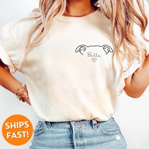 Pode incluir: Uma camiseta branca com um desenho simples de uma cabe&ccedil;a de cachorro e o nome "Bella" com um cora&ccedil;&atilde;o abaixo.