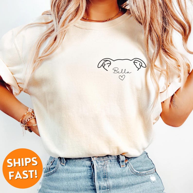 Pode incluir: Uma camiseta branca com um desenho simples de uma cabe&ccedil;a de cachorro e o nome "Bella" com um cora&ccedil;&atilde;o abaixo.