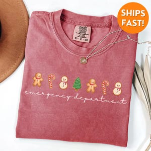 Puede incluir: Una camiseta roja con un gr&aacute;fico blanco de hombres de jengibre, bastones de caramelo, mu&ntilde;ecos de nieve y un &aacute;rbol de Navidad. El texto "emergency department" est&aacute; escrito debajo del gr&aacute;fico.