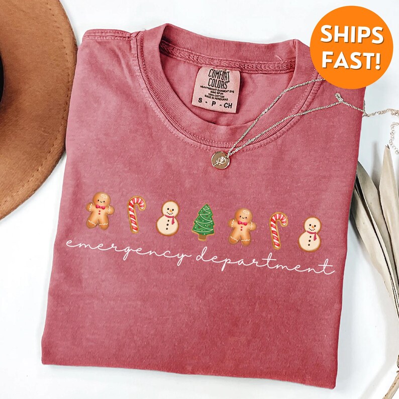 Puede incluir: Una camiseta roja con un gr&aacute;fico blanco de hombres de jengibre, bastones de caramelo, mu&ntilde;ecos de nieve y un &aacute;rbol de Navidad. El texto "emergency department" est&aacute; escrito debajo del gr&aacute;fico.
