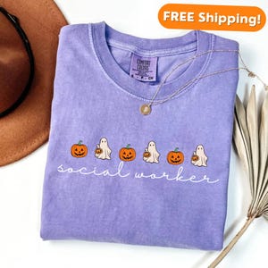 May include: A purple t-shirt with a white graphic of pumpkins and ghosts with the text "social worker" printed on the front.