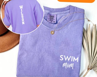 Puede incluir: Una camiseta de color lavanda con el texto "SWIM MOM" en la parte delantera y "WILSON" en la parte trasera. La camiseta está doblada y colocada sobre una superficie blanca.