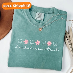 Pu&ograve; includere: Una maglietta verde acqua con una grafica bianca di denti e fiori rosa. Il testo "dental assistant" &egrave; scritto in corsivo bianco sotto la grafica.