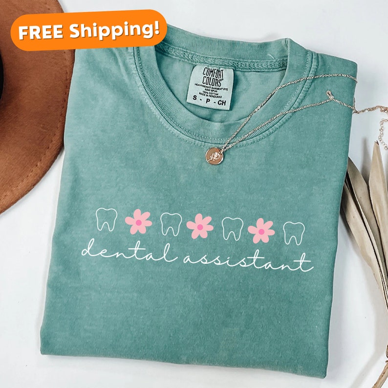 Pu&ograve; includere: Una maglietta verde acqua con una grafica bianca di denti e fiori rosa. Il testo "dental assistant" &egrave; scritto in corsivo bianco sotto la grafica.