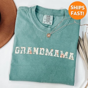 以下が含まれることがあります： 薄い青緑色のTシャツに、黒い縁取りで白く書かれた「GRANDMAMA」の文字。文字はピンク、白、緑の色の花柄で埋められています。Tシャツは使い古したような風合いがあり、柔らかく着心地の良い生地で作られています。