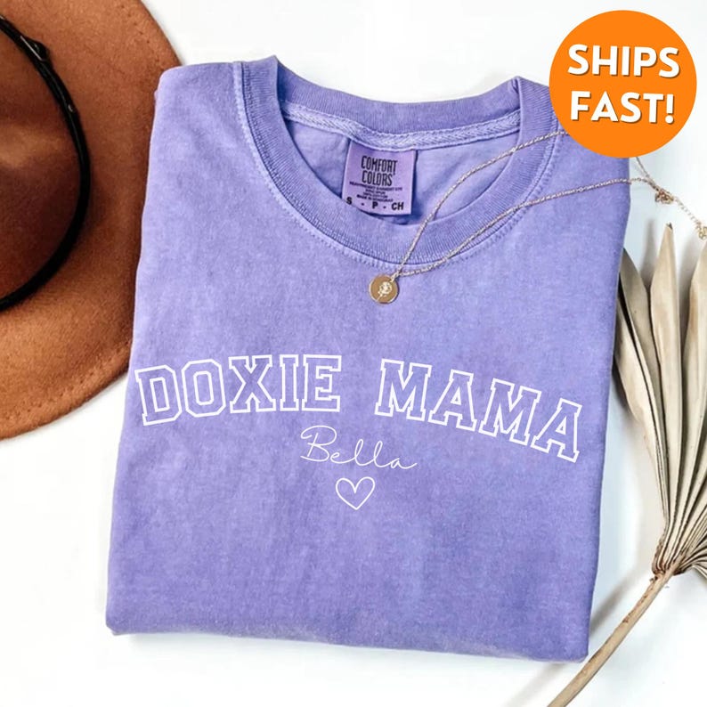 May include: Purple t-shirt with the text "DOXIE MAMA" in white outlined letters. The name "Bella" is written in cursive below the text with a heart.
