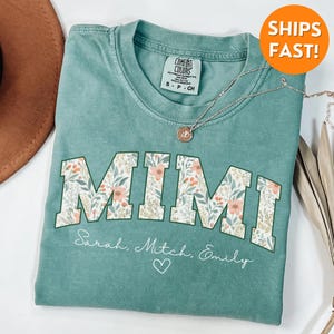May include: A teal green t-shirt with the word "MIMI" in white letters with a floral pattern. The text "Sarah, Mitch, Emily" is written below the word "MIMI" with a heart symbol.