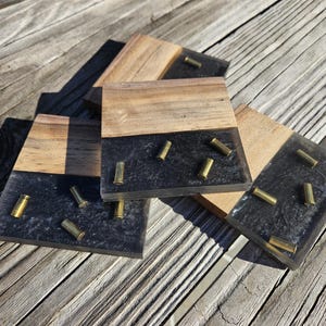 Puede incluir: Conjunto de cuatro posavasos hechos de madera y resina negra con casquillos de bala dorados incrustados en la resina.
