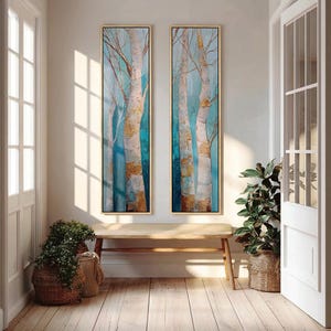May include: Two tall, vertical rectangular wall art pieces featuring a forest scene with white and gold birch tree trunks against a gradient blue background. Each painting is encased in a thin, light-colored wooden frame, displaying textured brushstrokes and a serene, nature-inspired composition.