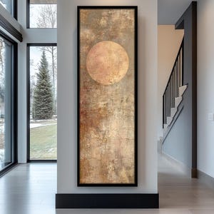 May include: A framed vertical abstract artwork. The piece features a large, textured, circular design in the upper centre, set against a background of mottled gold, brown, and beige tones. The artwork is displayed on a white wall.