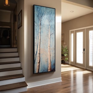 May include: A tall, vertical rectangular wall art piece featuring a forest scene with two prominent birch trees. The artwork is encased in a thin black frame and displays a soft blue and white color palette with light brown tree trunks.