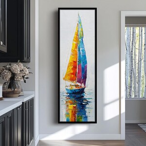 May include: A colorful sailboat painting with a black frame. The sailboat has a vibrant sail with yellow, orange, red, and blue hues. The water reflects the sail's colors. The artwork is displayed in a modern interior setting.