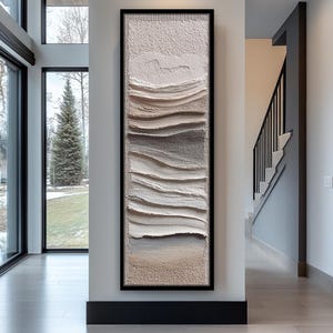 Pode incluir: Uma obra de arte abstrata vertical e longa em uma moldura preta. A obra apresenta camadas texturizadas de cores neutras, incluindo branco, bege e cinza, criando um padrão semelhante a ondas. É exibida em um corredor moderno.