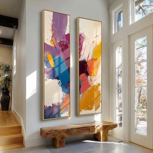 May include: Two vertical abstract paintings in gold frames. The artwork displays bold brushstrokes in shades of purple, blue, yellow, orange, and white. A wooden bench is positioned below the paintings, with a doorway and natural light.