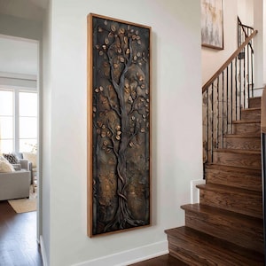 Può includere: Un'opera d'arte da parete alta e rettangolare che raffigura un albero scolpito in tre dimensioni con tronco e rami scuri e strutturati, decorati con foglie color bronzo metallizzato, il tutto racchiuso in una cornice di legno chiaro.