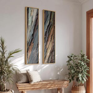 Op de afbeelding: Twee verticale ingelijste abstracte kunstwerken. Het kunstwerk heeft een textuur, veelkleurig ontwerp met tinten blauw, bruin en bruin. De frames zijn goudkleurig. De kunst wordt tentoongesteld op een witte muur boven een houten bank met kussens en planten.