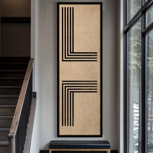 May include: A framed rectangular artwork with a beige background and black geometric lines. The black lines create angular shapes, forming an abstract design. The artwork is displayed in a hallway setting, above a black bench.
