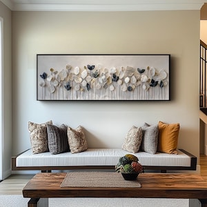 a living room with a large, rectangular artwork hanging on the wall above a white sofa with multiple pillows. The sofa is positioned in front of a wooden coffee table, which has a small vase of flowers on it. The overall scene has a modern and stylish aesthetic.