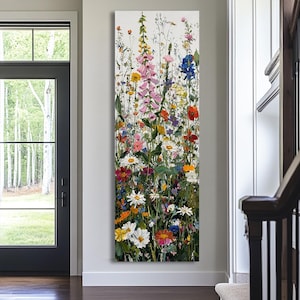 May include: A vertical canvas art piece featuring a vibrant wildflower meadow. The painting showcases a variety of colorful blooms, including pink foxgloves, daisies, and other wildflowers, set against a white background. The artwork is displayed in a hallway.