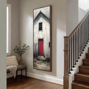 May include: Vertical framed art print of a white barn with a red door. The artwork has a rustic, textured appearance, with a dark wooden frame. The painting is displayed on a light gray wall in a hallway, near a staircase.