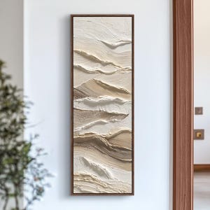 Puede incluir: Una obra de arte abstracta enmarcada de forma rectangular. La obra presenta pinceladas texturizadas en tonos blancos, beige y marrón, creando un efecto de capas. El marco marrón oscuro rodea la obra, que está montada en una pared blanca.