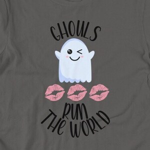 Könnte beinhalten: Ein dunkelgraues Kurzarm-T-Shirt mit einer Grafik eines zwinkernden weißen Geistes über drei rosa Lippenabdrücken. Der Text "GHOULS" ist in Schwarz über dem Geist und "RUN THE WORLD" in Schwarz unter den Lippenabdrücken aufgedruckt.