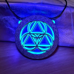 May include: A silver necklace with a pendant featuring a geometric design of overlapping triangles inside a circular frame with a Greek key border. The pendant glows green under a black light.