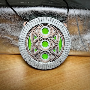 May include: A circular pendant necklace with a black cord. The pendant features a silver-colored outer ring with triangular cutouts, surrounding a copper-colored geometric design and a bright green center. The necklace is displayed on a wooden surface.