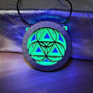 May include: A silver necklace with a round pendant featuring a green and blue geometric design. The pendant is surrounded by a decorative border.