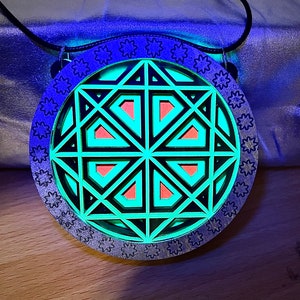 May include: A round, silver pendant with a black and green geometric design. The pendant has a black cord and is hanging from a silver chain.