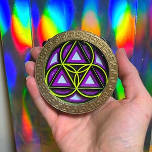 May include: A gold-colored metal pendant with a geometric design featuring overlapping purple, yellow, and black triangles. The pendant has a decorative border with a Greek key pattern.