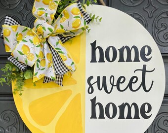 May include: A round wooden door sign with a lemon slice design and the words "home sweet home." The sign features a decorative bow with lemon and gingham patterns, and greenery. It hangs from a jute rope.