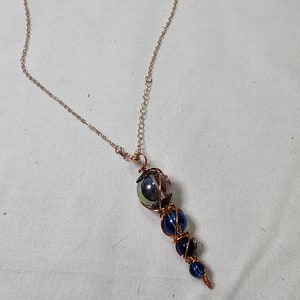 May include: A copper wire wrapped necklace with a blue and iridescent glass pendant. The pendant is made of multiple glass beads wrapped in copper wire.