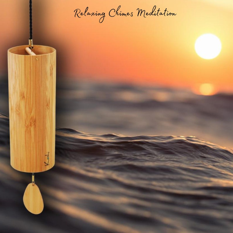 May include: A wooden wind chime with a natural finish and the word "Koti" inscribed on it. The chime is hanging from a black cord and is suspended over a blurry background of a sunset over the ocean.