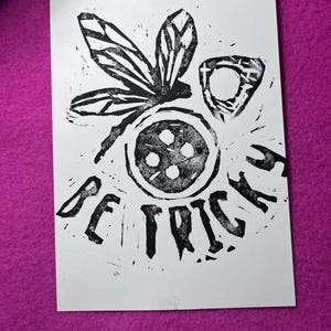 K&ouml;nnte beinhalten: Schwarz-Wei&szlig;-Linolschnitt auf wei&szlig;em Papier. Der Druck zeigt eine stilisierte Libelle, ein kreisf&ouml;rmiges Design mit vier Punkten und das Wort "BE TRICKED" in fetten Buchstaben. Das Kunstwerk ist vor einem magentafarbenen Hintergrund platziert.
