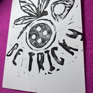 K&ouml;nnte beinhalten: Schwarz-Wei&szlig;-Linolschnitt auf wei&szlig;em Papier. Der Druck zeigt ein stilisiertes Insekt mit einem kreisf&ouml;rmigen Design und dem Wort "BEATRICE" in fetten Buchstaben. Der Druck befindet sich auf einem lila Hintergrund.