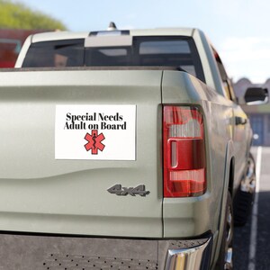 May include: A white rectangular bumper sticker with the text "Special Needs Adult on Board" in black. Below the text is a red medical symbol. The sticker is affixed to the back of a light green pickup truck.