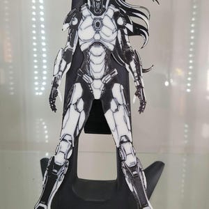 Peut inclure: Une figurine imprimée en 3D en blanc et noir d'une femme dans une combinaison futuriste. La femme a les cheveux longs et se tient debout, les jambes légèrement écartées. La figurine est sur un socle noir.