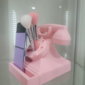 May include: A pink vintage-style phone shaped makeup brush holder with a purple phone case in the slot. Three makeup brushes are in the holder.