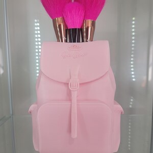 May include: Pink plastic backpack-shaped makeup brush holder with three pink makeup brushes sticking out of the top. The holder has the words "Girl Glam" embossed on the front.