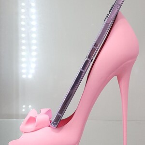 May include: A pink high-heeled shoe-shaped phone stand with a bow detail. A silver smartphone is resting in the stand. The stand is made of a solid, smooth material and has a stiletto heel.