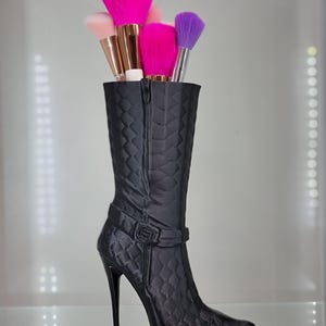 Könnte beinhalten: Ein schwarzer, gesteppter Stiefel mit hohem Absatz und spitzer Zehenkappe, der als Make-up-Pinselhalter verwendet wird. Der Stiefel ist mit rosa, lila und weißen Make-up-Pinseln gefüllt.