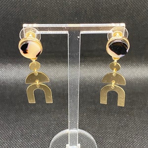 May include: Gold-toned dangle earrings with a geometric design. Each earring features a round stud with a black and tan marbled stone, connected to a series of gold shapes, including circles and half-moons. The earrings are displayed on a clear stand.