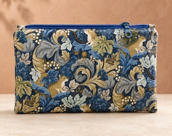 Può includere: Una pochette rettangolare in tessuto con cerniera, caratterizzata da un dettagliato motivo botanico con foglie blu e marrone chiaro, bacche bianche e piccoli scoiattoli marroni. La pochette presenta una chiusura a zip blu scuro.
