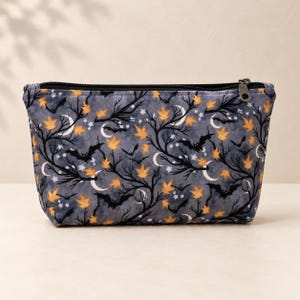 May include: A rectangular zippered pouch featuring a dark gray fabric with a repeating pattern of black silhouettes of bats, bare tree branches, white crescent moons, small white stars, and scattered orange autumn leaves throughout the design.