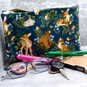 May include: A dark teal zippered pouch with a woodland animal print featuring deer, rabbits, foxes, and birds. The pouch has a gold zipper and is next to a green pen, glasses, keys, and a phone.