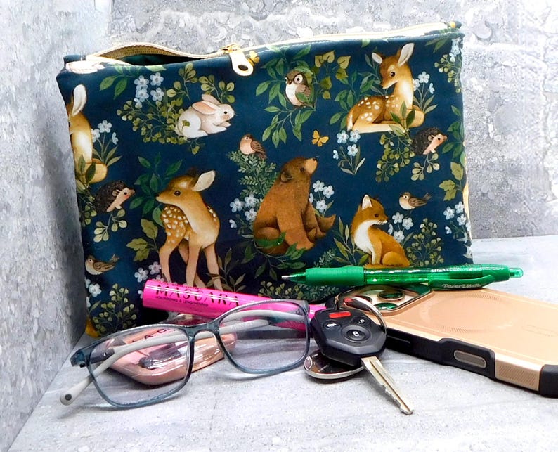 May include: A dark teal zippered pouch with a woodland animal print featuring deer, rabbits, foxes, and birds. The pouch has a gold zipper and is next to a green pen, glasses, keys, and a phone.