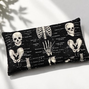 May include: Black rectangular pillow featuring a detailed anatomical illustration of the human skeletal system. The design includes skulls, rib cages, and bone diagrams in white against a black background. The pillow is a decorative item.